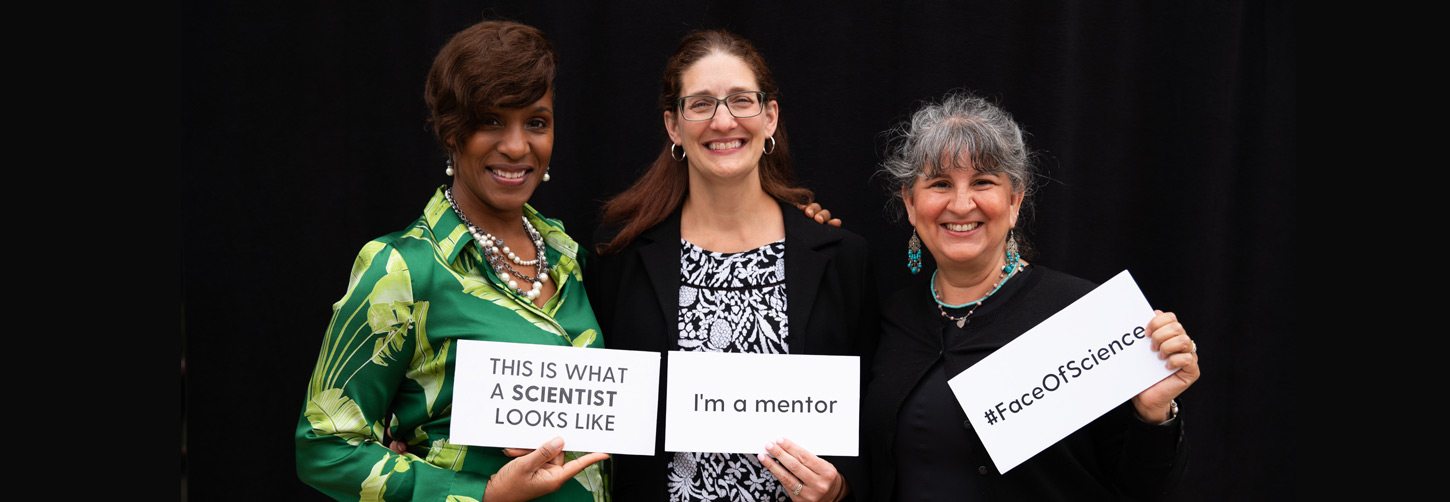 Three people who are each holding a sign. Each sign says: This is what a scientist looks like, I'm a mentor, and hashtag FaceOfScience.